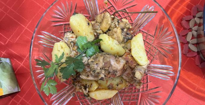 Fotografia vista de cima de um recipiente fundo de vidro com frango cozido, batatas e coentro por cima. O recipiente está sobre uma toalha de mesa vermelha.