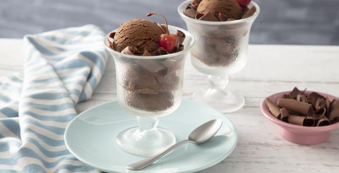 Fotografia em tons de azul de uma bancada branca, ao centro um pratinho azul com uma taça com duas bolas de sorvete de chocolate e uma cereja. Ao fundo outra taça com sorvete de chocolate e um potinho com raspas de chocolate.