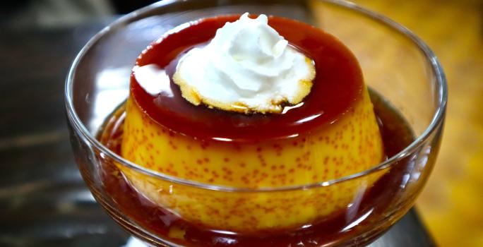 Fotografia de um mini pudim de café dentro de uma taça pequena de vidro com a calda de caramelo. Por cima do pudim tem um pouco de chantilly.