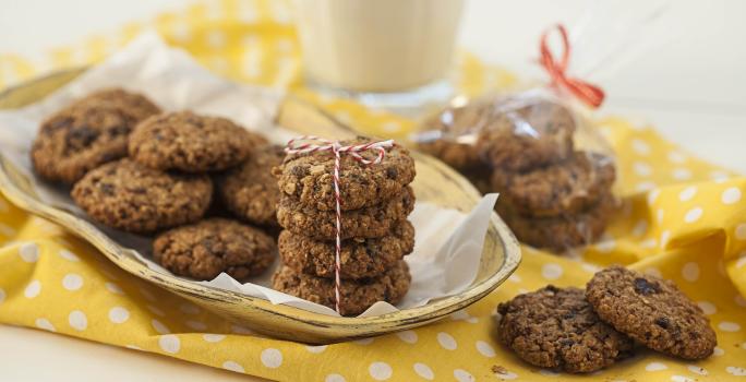 Fotografia em tons de branco e amarelo de uma bancada branca com um paninho amarelo com bolinhas brancas, sobre ele cookies. Ao fundo um copo de vidro.