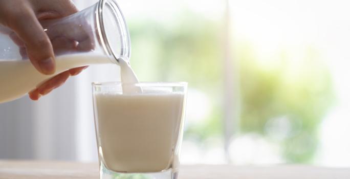 Fotografia de uma mão colocando uma bebida de iogurte com leite e cereal em um copo de vidro, que está sobre uma mesa de madeira em tom claro.