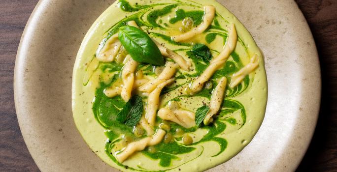 Fotografia em tons de cinza com um prato cinza e fundo ao centro. Dentro do prato existe uma receita de macarrão trofie com molho de abobrinha e ervilhas coberto com pesto de ervas.