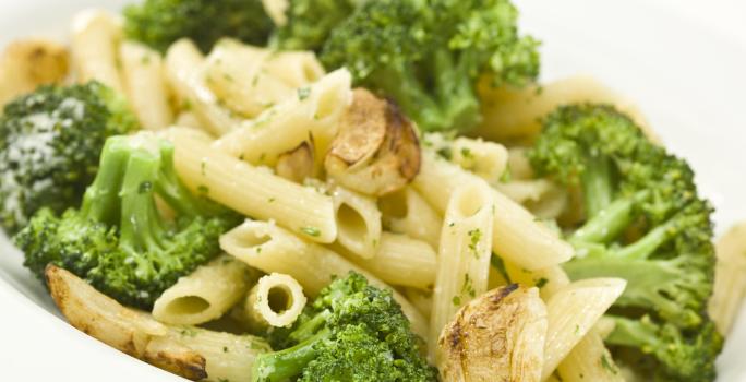 Fotografia de uma receita de penne com brócolis, um pouco de azeitona e alho assado dentro de um refratário fundo branco sobre uma mesa de cor branca.