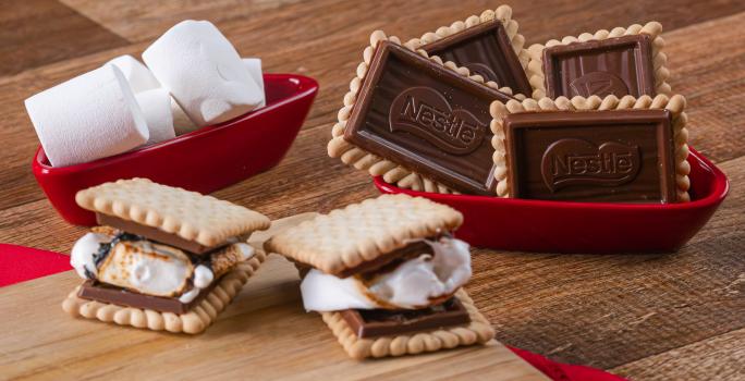 Fotografia em tons de vermelho com vários recipientes vermelhos ao centro. Dentro de um dos recipientes, existe dois biscoitos Choco Biscuit recheados com marsmallow. Ao lado existe marshmallows e biscoitos Choco Biscuit NESTLÉ