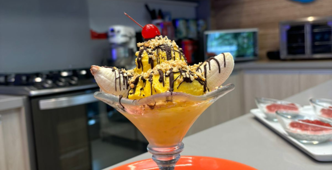 Fotografia de uma taça de vidro com banana split com calda de chocolate, farofa de castanha e sorvete de abacate.