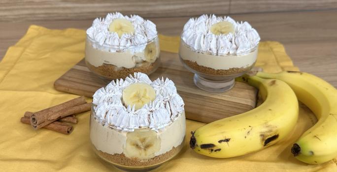 Fotografia em tons de amarelo com um pano amarelo ao centro. Em cima do pano existe uma receita de banoffee feita em pequenos potes de vidro cobertos com chantilly. Ao lado existe duas bananas.