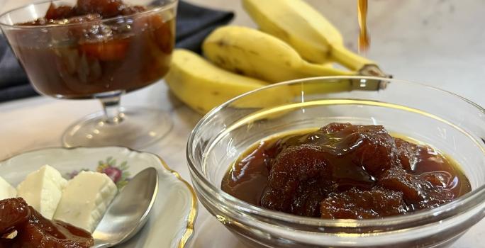 Foto da receita de compota de banana servida uma parte em um bowl de vidro e outra em uma taça baixa atrás. Ao lado, há um prato de porcelana com queijo branco e compota de banana e uma colher prateada. Ao fundo um cacho de bananas e um pano preto.