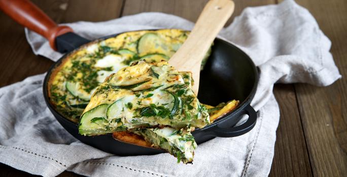 Foto da receita de omelete de abobrinha, servida em uma frigideira preta de ferro, com uma fatia mais próxima, sobre uma colher de pau. A frigideira está sobre um pano bege em uma bancada de madeira.