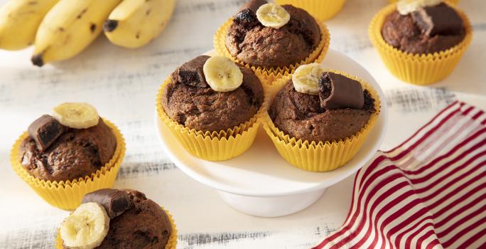 Foto da receita de Muffin de Banana com Chocolate Caribe. Observa-se 8 muffins em forminhas amarelas com uma rodela de banana e metade de um bombom no topo.