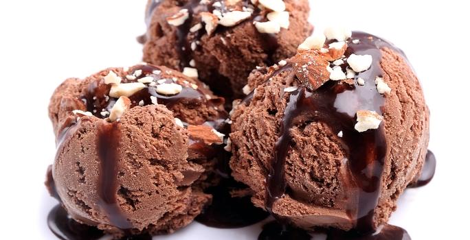 Fotografia de três bolas de sorvete de chocolate com cobertura de chocolate e farofa doce. A sobremesa está em um fundo branco.