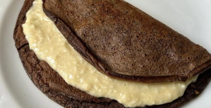 Fotografia de uma panqueca de chocolate com recheio de coco cremoso sobre um prato branco de vidro.