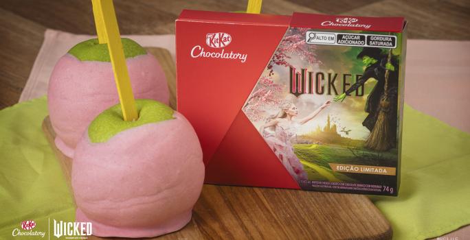 Foto da receita de Maçã verde com cobertura rosa de chocolate. Elas estão sobre uma bancada de madeira com um tecido verde e rosa e uma caixa de KitKat Wicked à direita