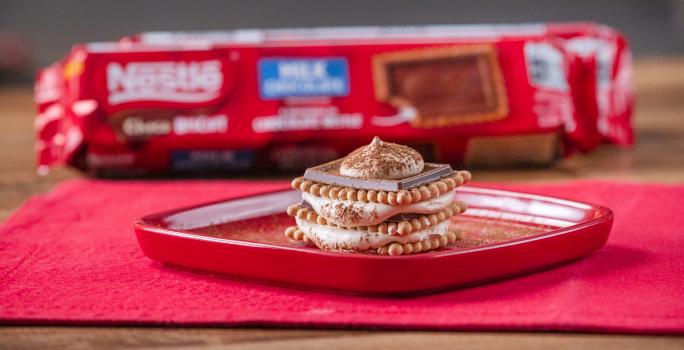 Fotografia de um tiramissu em camadas de creme e choco biscuit sobre um prato quadrado vermelho e raso com cacau em pó polvilhado. O prato está apoiado em um pano vermelho, e ao fundo, tem um pacote de choco biscuit.