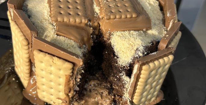 Fotografia de um bolo de chocolate com brigadeiro e farinha láctea polvilhada. Ao redor do bolo tem chocotrio de amendoim grudado. O bolo está sobre um prato de vidro preto raso.