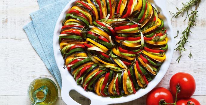 Fotografia em tons de verde, amarelo e vermelho de uma bancada branca com três tomates, uma jarrinha de azeite, ramos de alecrim e um recipiente branco redondo com alças com camadas intercaladas de abobrinha, berinjela e pimentões.