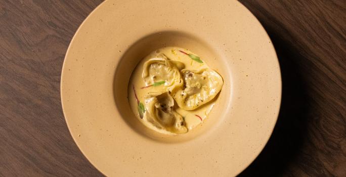 Foto da receita de Ravioli de Escargot feito no Masterchef 11. Observa-se um prato fundo visto de cima com três raviolis servidos com molho e decorado com brotos.