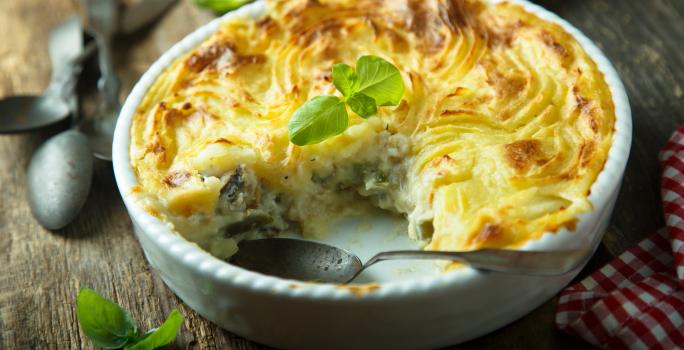 Foto em tons de amarelo da receita de torta de bacalhau servida em uma forma de porcelana branca com uma fatia cortada separada da torta e uma colher prateada dentro