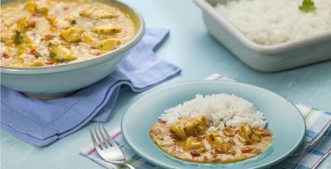 Fotografia em tons de azul e amarelo, ao centro prato azul com frango ensopado e arroz, sobre guardanapo azul e branco com um garfo, à direita travessa com arroz, à esquerda travessa com frango ensopado e guardanapo azul, tudo sobre bancada na cor azul.