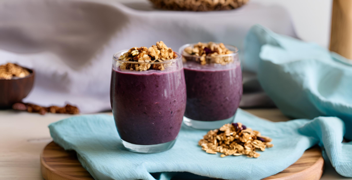 Fotografia de dois copos de vidro, os dois com shake de açaí e granola por cima. Os copos estão sobre um pano azul de tom claro, com mais granola, no canto. O pano está sobre uma tábua redonda de madeira.