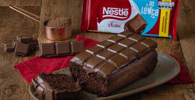 Foto da receita de Chocolate Bar Cake, sobre uma bancada de madeira, em um prato em tom marrom. Na bancada há uma barra de chocolate ao leite, um copo de medida de cobre com cacau em pó e alguns pedaços de chocolate