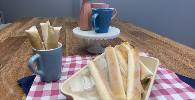 Foto da receita de Sacolé de Pudim de Leite Moça, servido em saquinhos com caramelo, sobre uma travessa com cubos de gelo, numa bancada de madeira decorada com um tecido rosa e branco e com canecas e um vaso cor de rosa