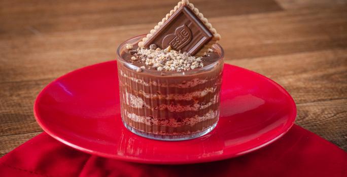 Fotografia de um copo de vidro pequeno com camadas de chocolate alpino derretido e chocobiscuit triturado. Por cima tem choco biscuit triturado e um biscoito inteiro apoiado para decorar. O copo está sobre um prato e uma toalha de mesa vermelha.