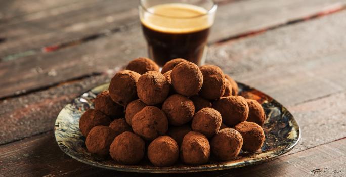 fotografia em tons de marrom e preto de uma bancada marrom vista de frente, contém um prato redondo com trufas com chocolate, e ao fundo um copo com café.