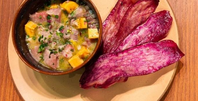 Fotografia vista de cima de um recipiente pequeno com ceviche ao lado de 4 lâminas de batata doce frita, sobre um prato de tom claro apoiado em uma bancada de madeira.