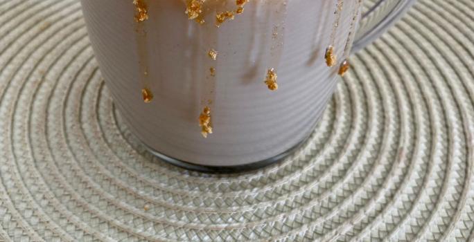 Fotografia em tons de marrom em uma bancada com um pano bege e uma xícara de vidro com a bebida de cappuccino de caramelo salgado dentro. Xícara decorada com calda de caramelo e biscoitos Tostines Especiarias