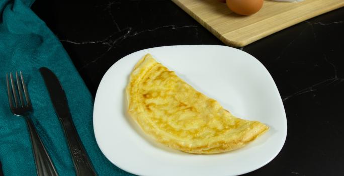 Fotografia em tons mais escuros de um prato branco com uma crepioca dobrada na metade. O prato está sobre uma mesa preta e ao lado de talheres, os quais estão apoiados em um pano de tom azul.