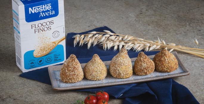 Foto da Receita de Coxinha da Batata Doce com Nestlé Aveia Flocos Finos, servidas em um prato cinza retangular, ao todo são 5, numa bancada de pedra com um tecido azul escuro, uma caixa de nestlé aveia flocos finos.