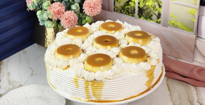 Foto da Receita de Bolo Recheado de Pudim com Doce de Leite Moça, decorado sobre um prato branco com sete mini pudins por cima, em uma bancada de mármore com flores ao fundo