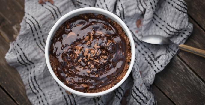 Fotografia vista de cima de um baked oats de air fryer dentro de um ramekin branco apoiado em um pano de tom cinza amassado, o qual está sobre uma madeira escura. Sobre o bolo tem uma calda de chocolate.