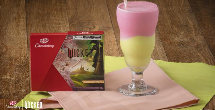 Foto aproximada de um copo alto com a receita de milkshake dividido em cores verde e rosa, servido sobre uma bancada de madeira com tecidos verde e rosa e com a caixa de KitKat Wicked ao lado esquerdo