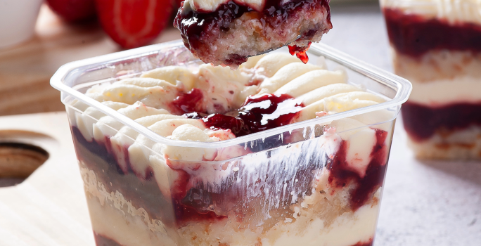 Fotografia mostra um bolo recipiente transparente mostrando camadas de um bolo de pote com creme branco e geléia de frutas vermelhas.