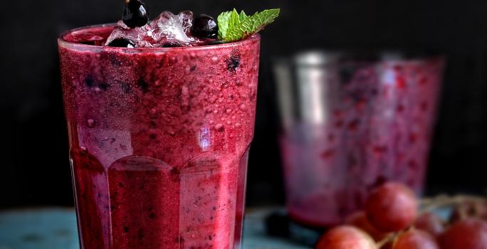 Fotografia em tom escuro de dois copos de vidro, o da frente está cheio com uma bebida de suco de uva com leite e neston, e o do fundo está quase vazio.