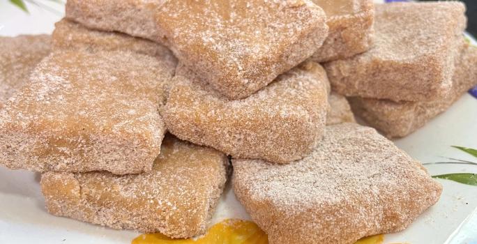 Fotografia de vários quadradinhos de palha italiana de churros, passados no açúcar e canela. As unidades estão juntas e sobrepostas, apoiadas em um prato branco com desenhos coloridos