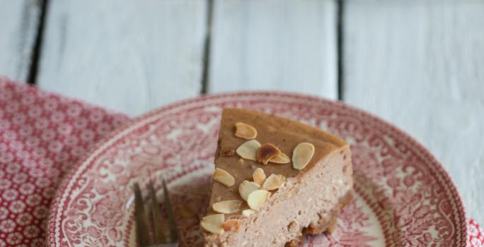 Fotografia de um prato de vidro desenhado com cor vermelho e branco. Sobre esse prato tem uma fatia de uma cheesecake de chocolate com amêndoas, ao lado de um garfo. Por cima da sobremesa tem lâminas de amêndoa. O prato está sobre um pano da mesma cor.