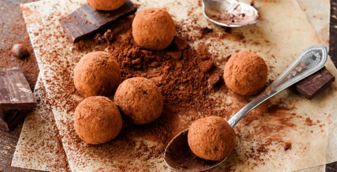 Fotografia vista de cima de algumas trufas de café e chocolate passadas no cacau em pó, sobre uma tábua de madeira salpicada com cacau em pó. Tem alguns pedaços de chocolate em volta das trufas e uma colher segurando uma delas.