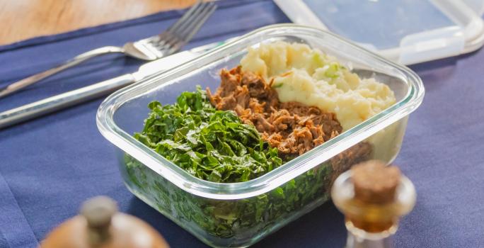 A fotografia mostra uma marmita em pote de vidro com carne desfiada ao meio, purê de batata-doce cremoso no canto superior direito e couve refogada verde no canto esquerdo.