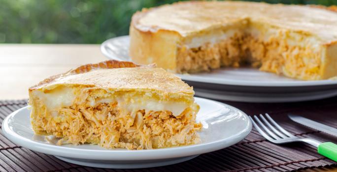 Fotografia de uma fatia de torta de frango com requeijão sobre um prato pequeno de vidro raso. No fundo tem um apoio branco e preto com o restante da torta quase inteira.