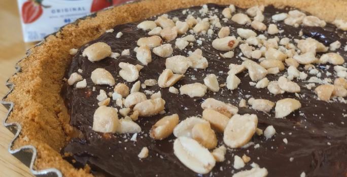 Fotografia de uma torta inteira charge ainda dentro da forma de alumínio. Por cima da torta tem amendoim, no fundo tem creme de leite e chocolate. A forma está sobre uma tábua de madeira.