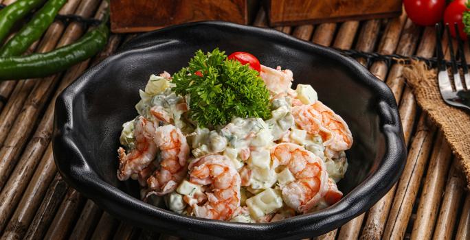 Fotografia de um recipiente preto fundo com uma salada de maionese com batata e camarão, com um tomate cereja e ramos de salsinha por cima. O recipiente está sobre uma mesa com madeiras desenhadas.