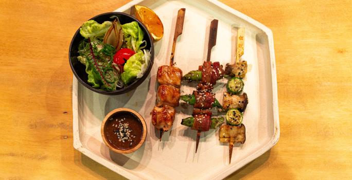 Fotografia vista de cima de um recipiente branco em formato de hexágono sobre uma mesa de madeira. Dentro desse recipiente tem um pote preto pequeno com salada, ao lado tem três espetinhos japonês com frango, bacon e legumes, e um molho ao lado.