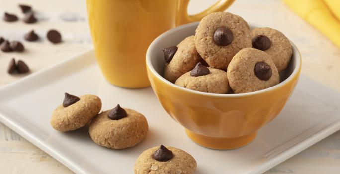 Fotografia em tons de branco e amarelo de uma bancada branca com um prato quadrado, sobre ele uma xícara amarela, um recipiente redondo amarelo com biscoitinhos  e três biscoitinhos. Ao fundo um paninho amarelo e gotas de chocolate.