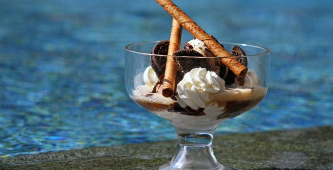 Fotografia de uma taça grande decorativa com cassata de chantilly e sorvete de Chocolate com alguns biscoitos e dois tubinhos de chocolate. O recipiente está apoiado sobre a base de uma piscina.