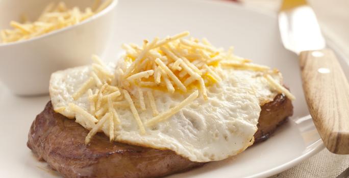 fotografia em tons de rosa, branco e marrom de uma bancada rosa vista de frente, contém um prato redondo branco com um pedaço de carne com ovo por cima e batata palha. Ao lado garfo e faca para servir, e ao fundo uma jarra e um copo transparente com suco.