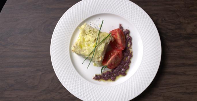 Fotografia em tons de branco com um prato branco ao centro. Dentro do prato existe uma receita de bacalhau acompanhada por tomates e azeitonas