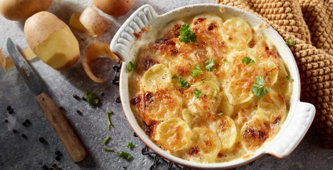 Foto da receita de Batata Gratinada Zero lactose. Observa-se um refratário redondo branco de porcelana com as batatas gratinadas dentro. Ao lado esquerdo, batatas descascadas decoram