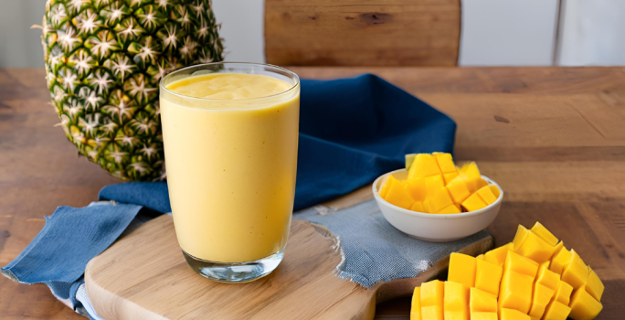 Fotografia de um copo de vidro com smoothie de iogurte, manga e abacaxi. O copo está sobre uma mini tábua de madeira, ao lado de dois recipientes brancos pequenos com pedaços de manga. No fundo tem um abacaxi inteiro sobre um pano azul escuro.